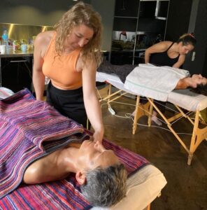 Zdjęcie przedstawia naukę masażu Peloha. Dwie uczestniczki same uczą się na swoich koleżankach z kursu, które leżą na stołach do masażu.